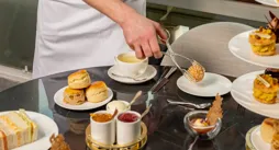 Chef plating pastries and tea at table with scones, sandwiches, and desserts, smiling beside window in bright dining setting.