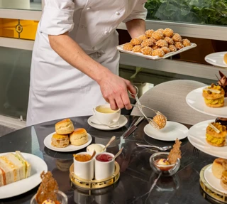 Chef plating pastries and tea at table with scones, sandwiches, and desserts, smiling beside window in bright dining setting.