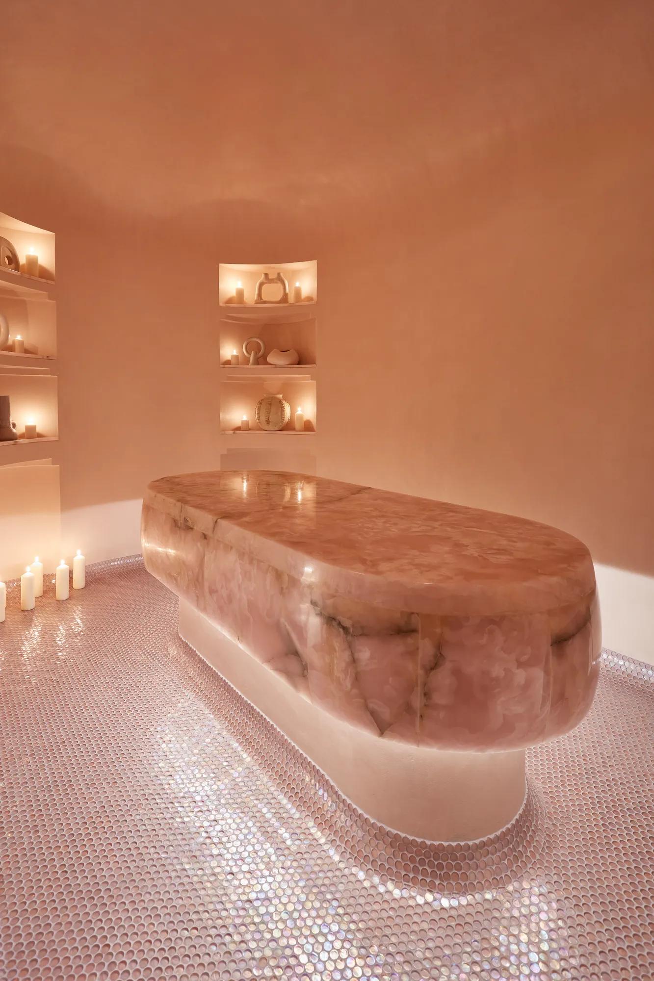 Softly lit spa room with a sculptural stone treatment table, candlelight, mosaic floor, and recessed shelves with ceramics.