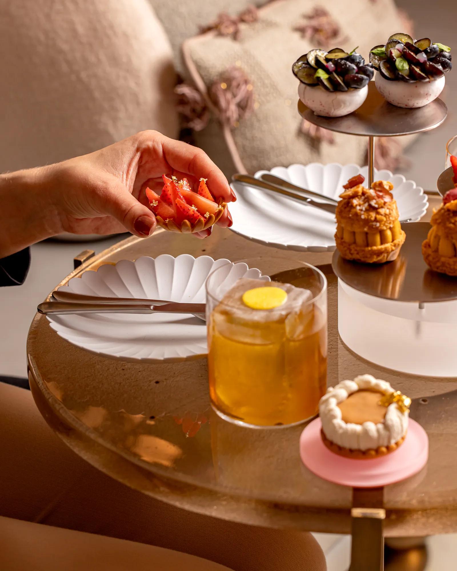 Hand holding a strawberry tart beside cocktails and elegant savoury pastries on a marble table during afternoon tea.