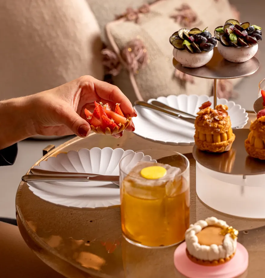 Hand holding a strawberry tart beside cocktails and elegant savoury pastries on a marble table during afternoon tea.