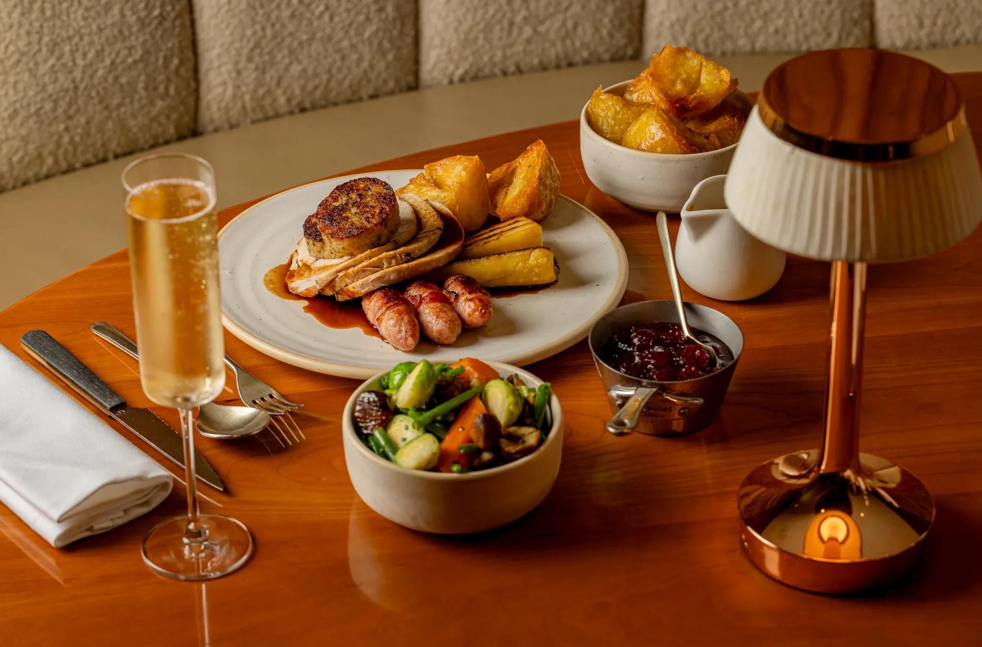 Roast dinner with turkey, stuffing, sausages, potatoes, vegetables, gravy and a glass of sparkling wine on a table.