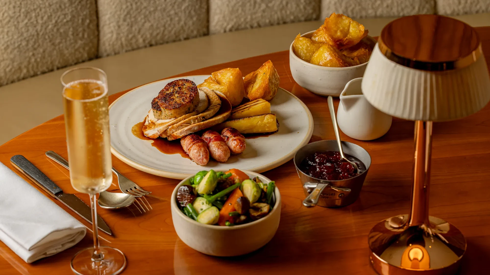 Roast dinner with turkey, stuffing, sausages, potatoes, vegetables, gravy and a glass of sparkling wine on a table.