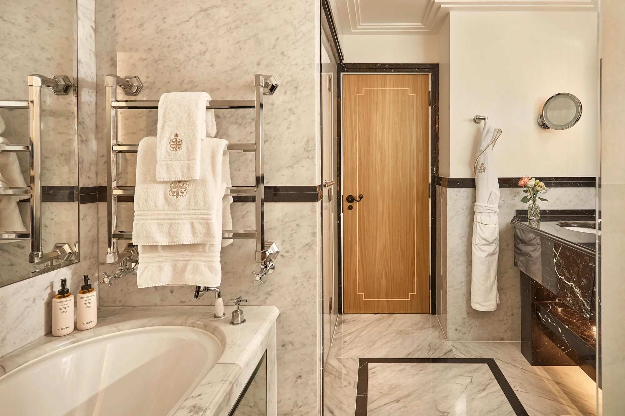 Elegant marble bathroom featuring a bathtub with branded towels on a heated rack, double vanity with dark marble accents, and a wooden door at the back, softly lit with warm lighting.