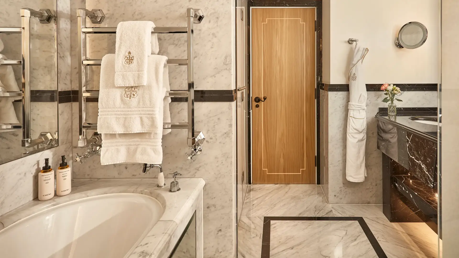 Elegant marble bathroom featuring a bathtub with branded towels on a heated rack, double vanity with dark marble accents, and a wooden door at the back, softly lit with warm lighting.