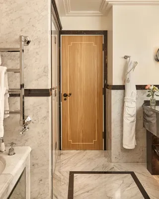 Elegant marble bathroom featuring a bathtub with branded towels on a heated rack, double vanity with dark marble accents, and a wooden door at the back, softly lit with warm lighting.