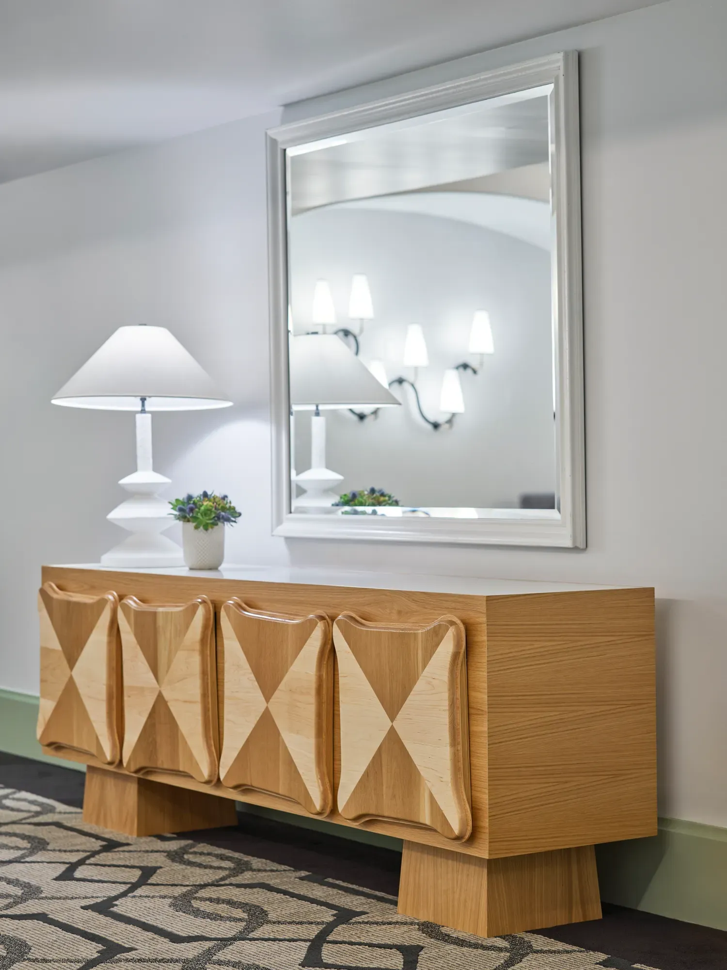 Gallery foyer vignette: sculpted wood credenza, white table lamp, and large mirror reflecting wall sconces.