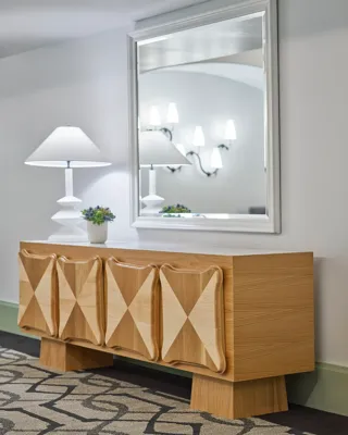 Gallery foyer vignette: sculpted wood credenza, white table lamp, and large mirror reflecting wall sconces.