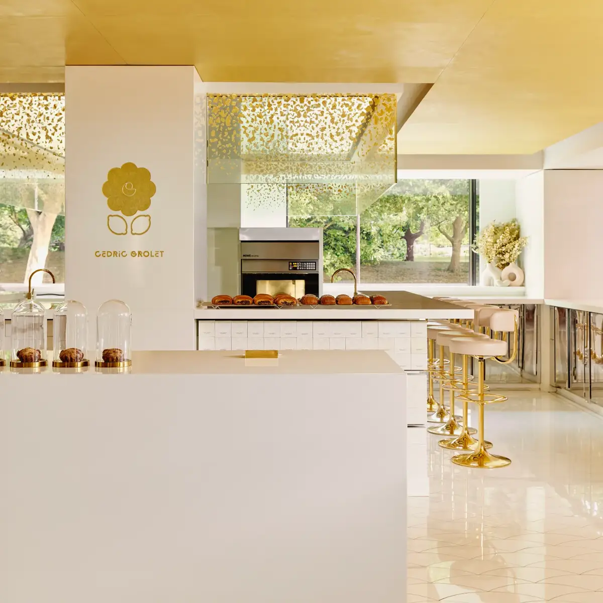 Bright patisserie interior with gold accents, marble counter, and pastries displayed beneath glass cloches.