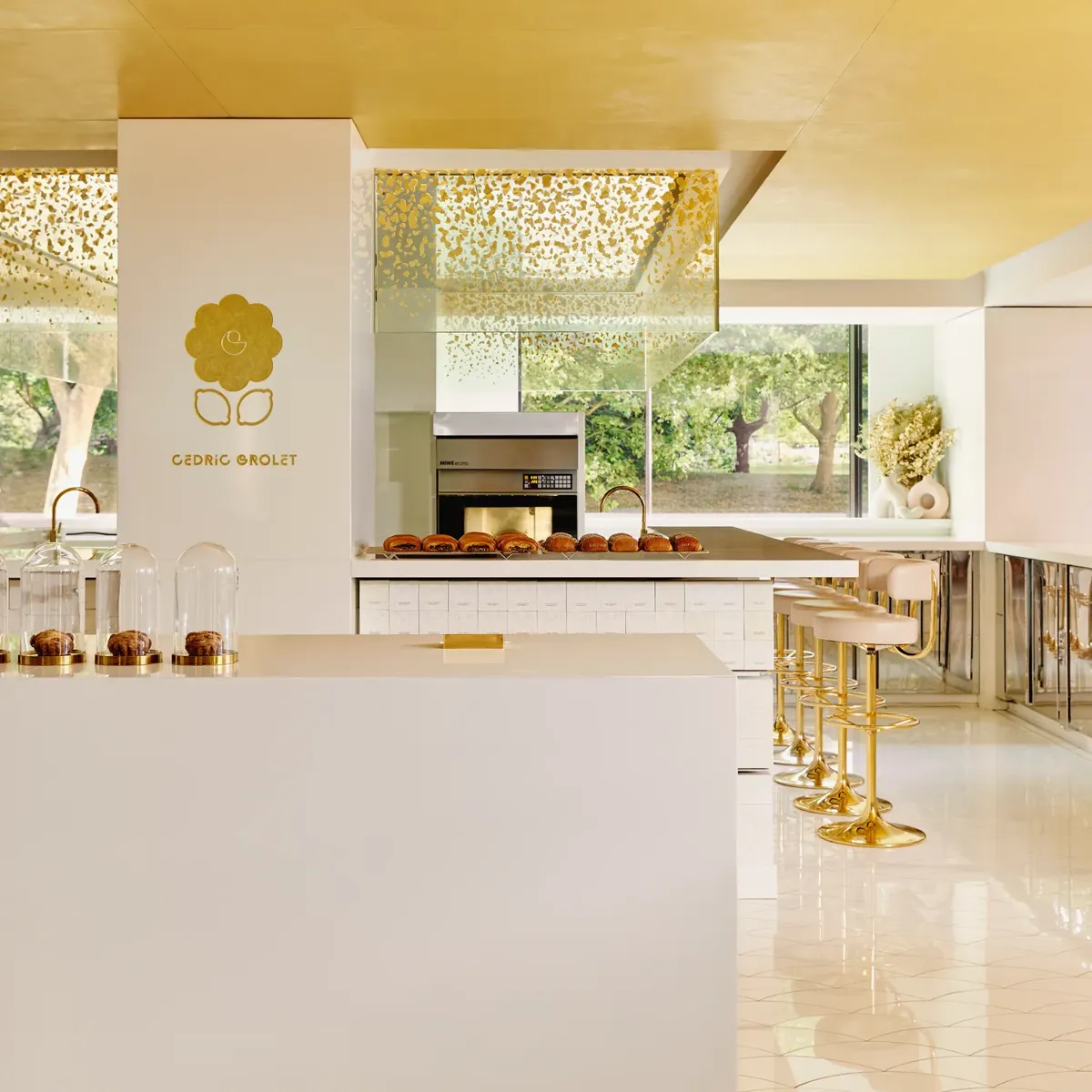 Bright patisserie interior with gold accents, marble counter, and pastries displayed beneath glass cloches.