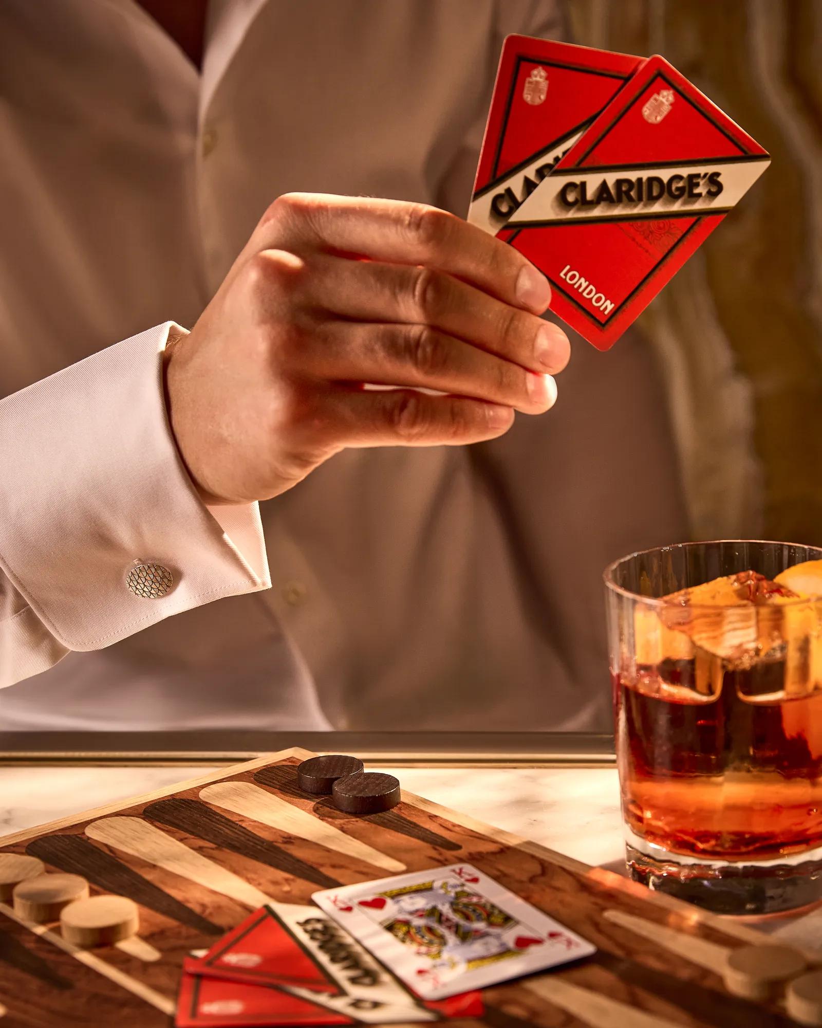 Hand holding Claridge’s London playing cards above backgammon board, with whiskey glass and pieces on table.