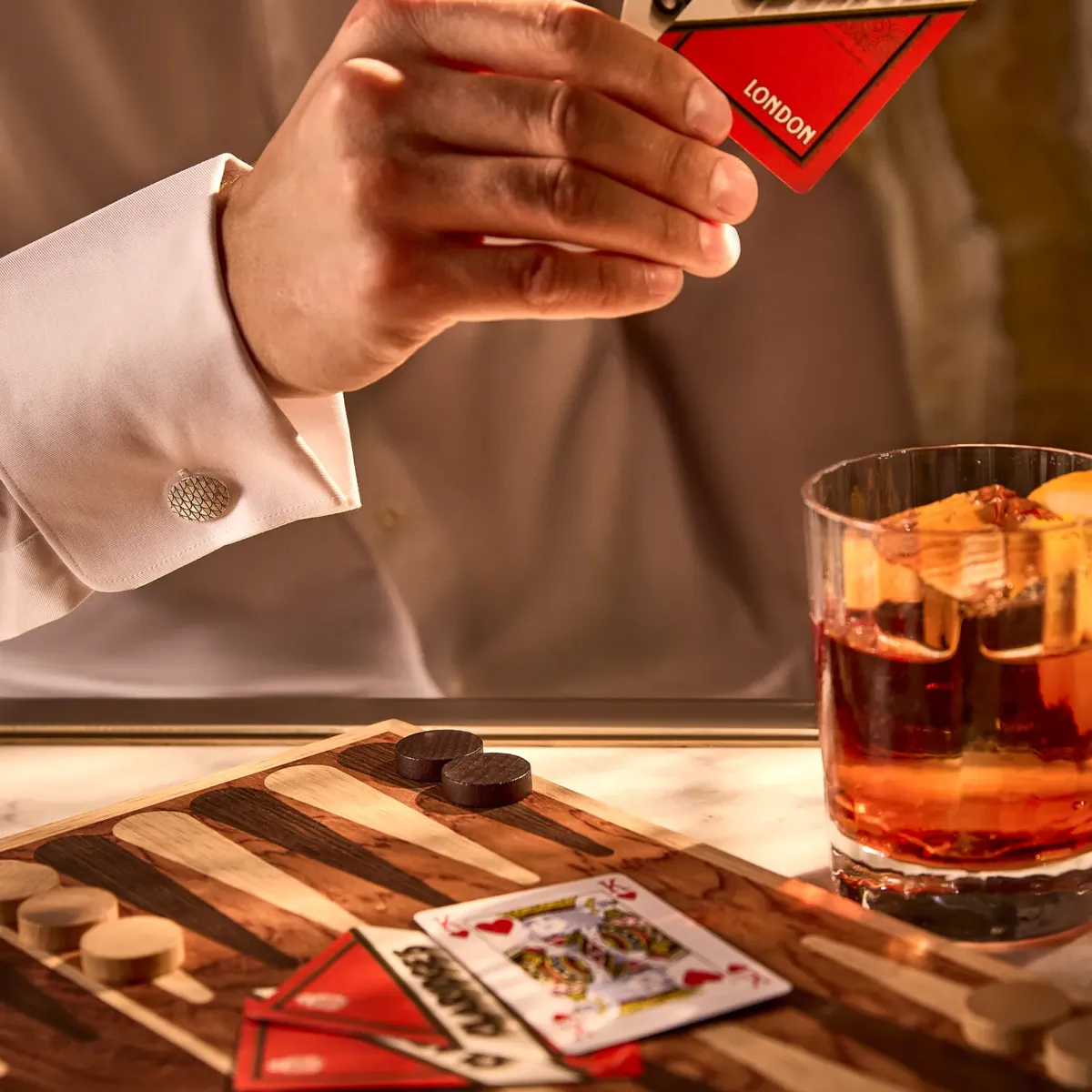 Hand holding Claridge’s London playing cards above backgammon board, with whiskey glass and pieces on table.