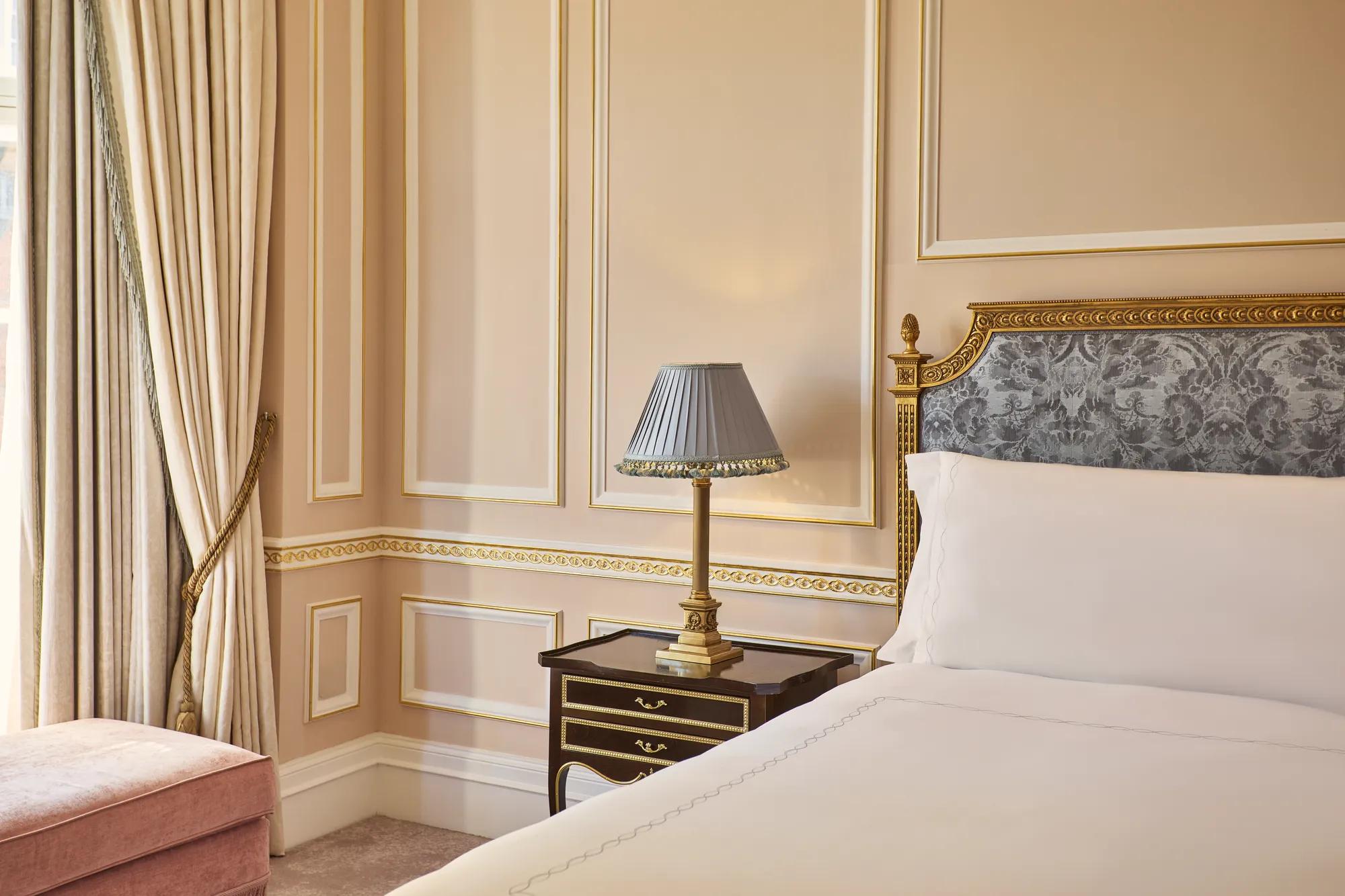 Partial view of a king-size bed with an upholstered headboard, bedside table and lamp, set against blush-pink walls with gold detailing and floor-length curtains.