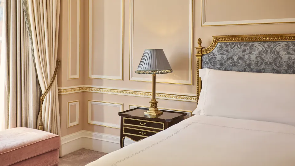 Partial view of a king-size bed with an upholstered headboard, bedside table and lamp, set against blush-pink walls with gold detailing and floor-length curtains.