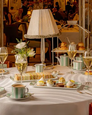 Elegant afternoon tea setting with a round table laid with striped china, pastries, finger sandwiches, and champagne glasses. A soft-lit table lamp and floral arrangement create a warm, refined atmosphere in a lively dining room.