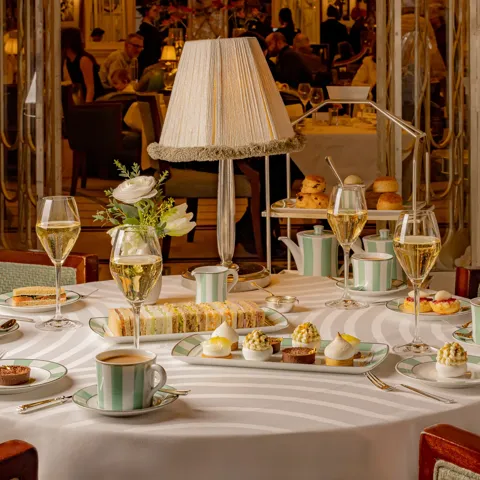 Elegant afternoon tea setting with a round table laid with striped china, pastries, finger sandwiches, and champagne glasses. A soft-lit table lamp and floral arrangement create a warm, refined atmosphere in a lively dining room.