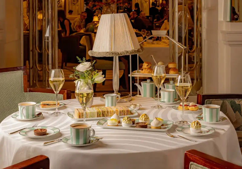 Elegant afternoon tea setting with a round table laid with striped china, pastries, finger sandwiches, and champagne glasses. A soft-lit table lamp and floral arrangement create a warm, refined atmosphere in a lively dining room.