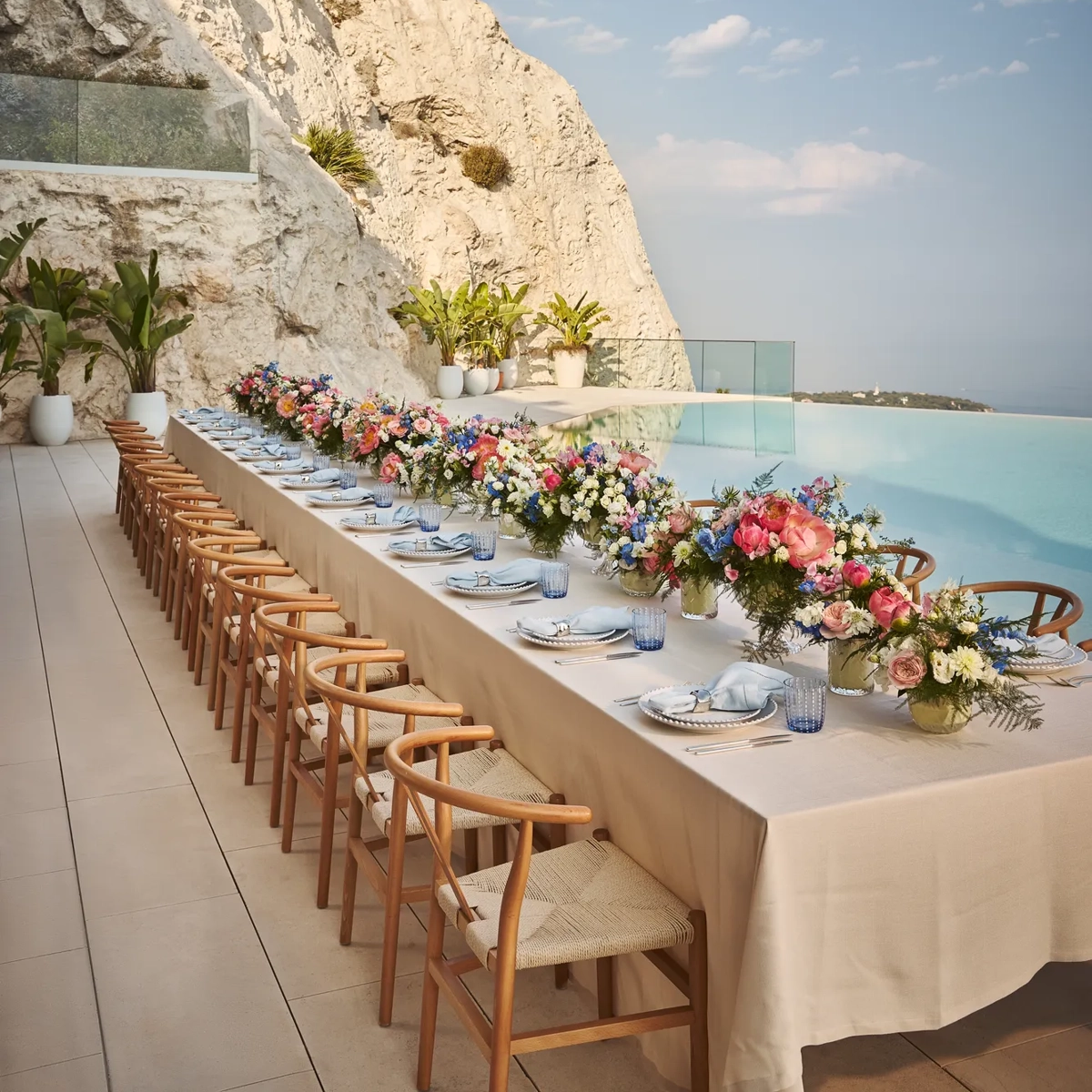 Longue table de repas en extérieur avec nappe crème, chaises en bois et compositions florales colorées, installée au bord d’une piscine à débordement au pied d’une falaise rocheuse.
