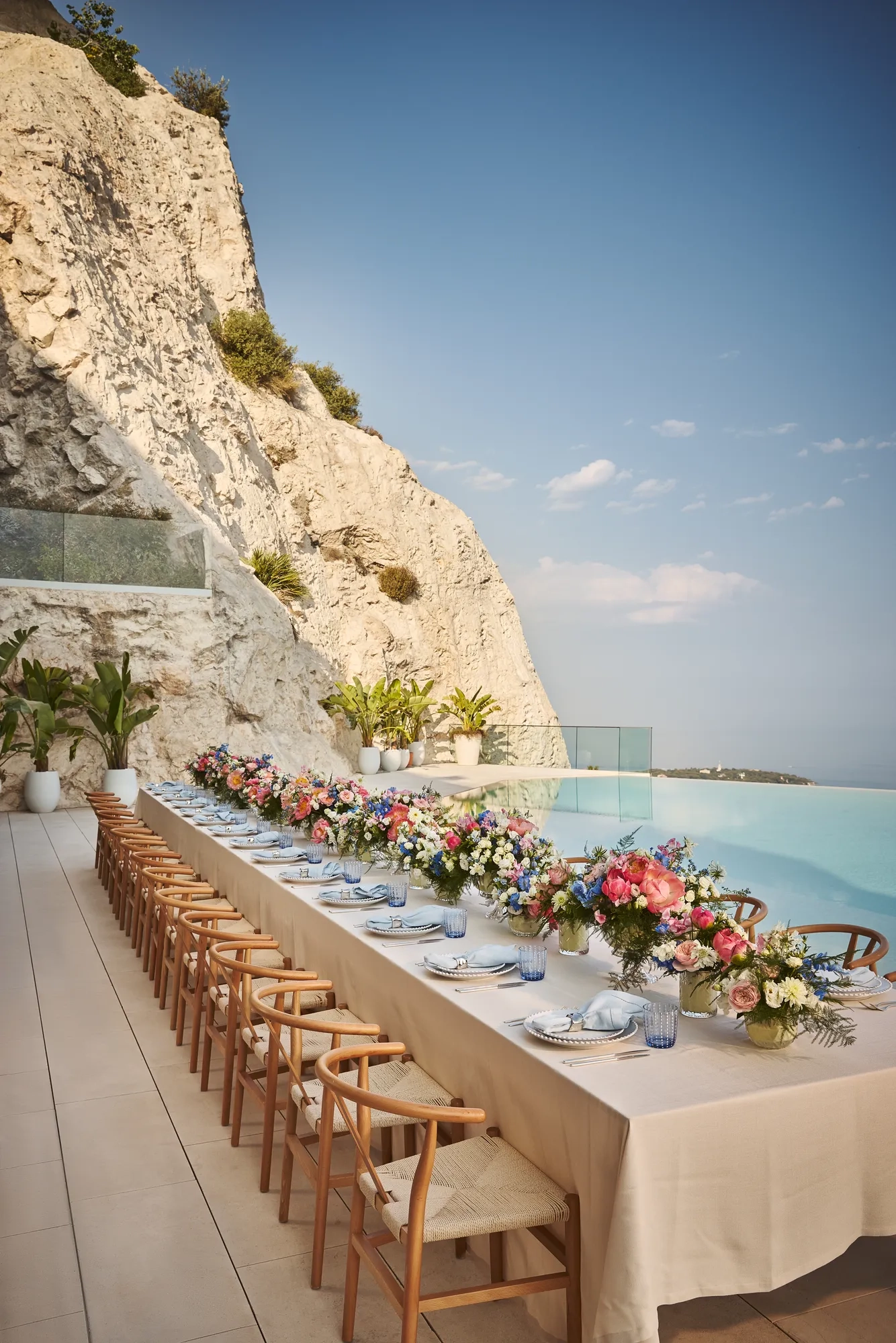 Longue table de repas en extérieur avec nappe crème, chaises en bois et compositions florales colorées, installée au bord d’une piscine à débordement au pied d’une falaise rocheuse.