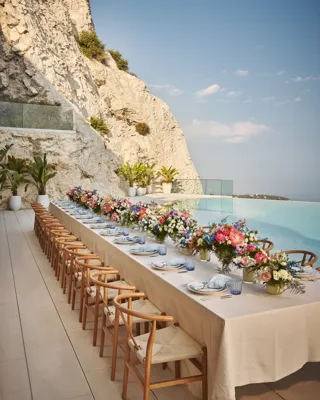 Longue table de repas en extérieur avec nappe crème, chaises en bois et compositions florales colorées, installée au bord d’une piscine à débordement au pied d’une falaise rocheuse.