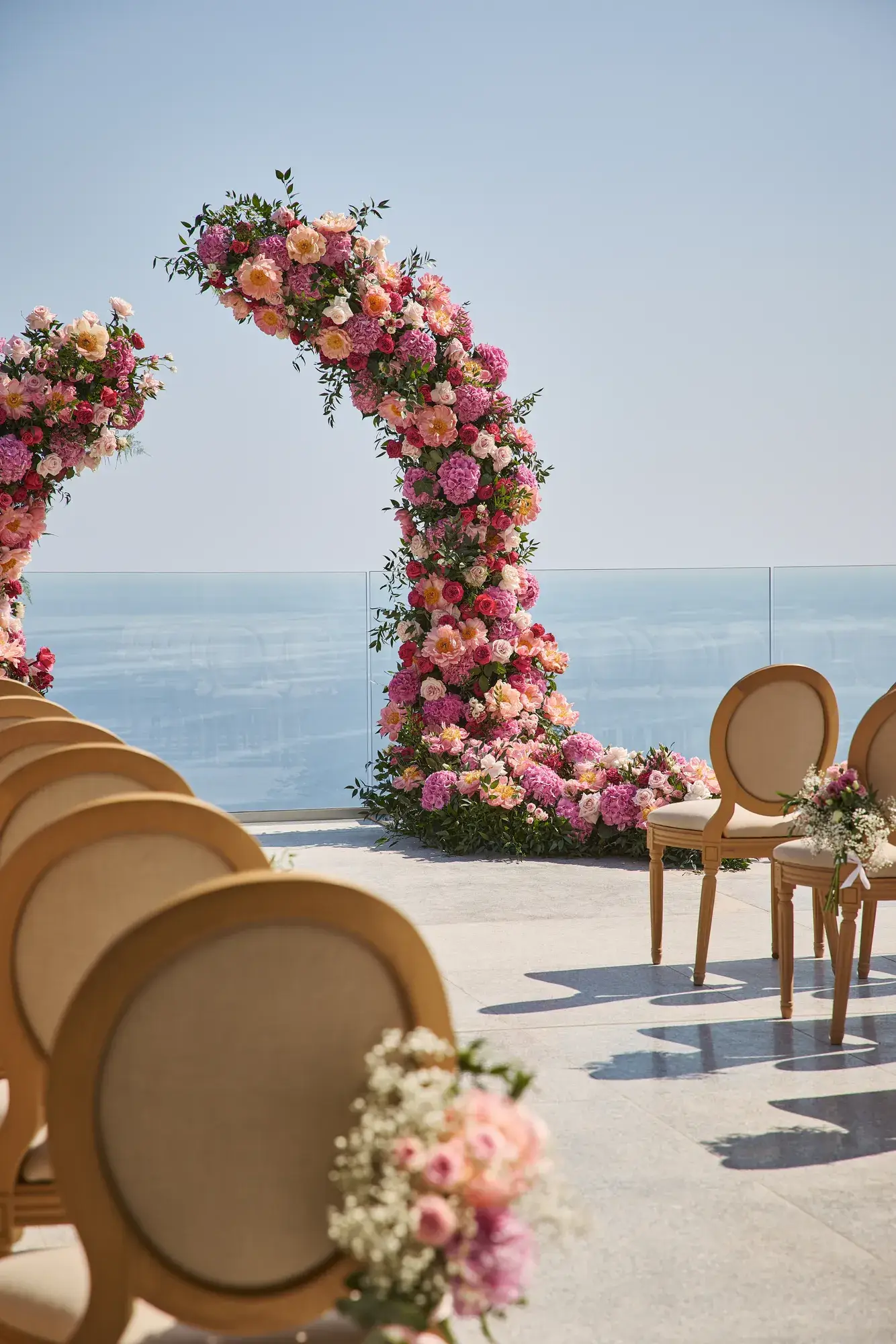 Arche de mariage florale aux fleurs roses et pêches en bord de mer, avec chaises décorées pour la cérémonie.