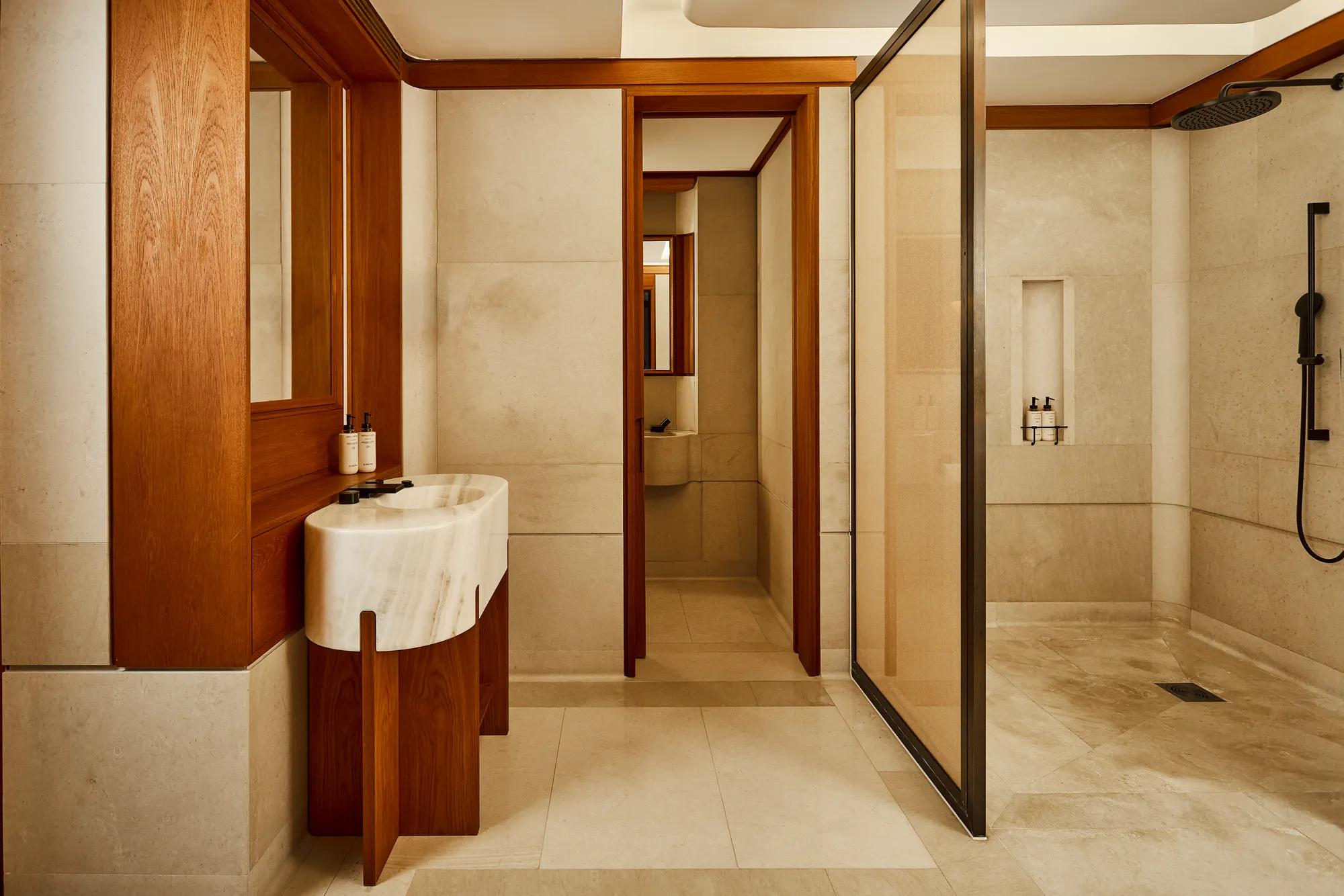 Bathroom with a marble vanity, walk-in rainfall shower, and warm wood detailing in a spacious stone-clad interior.
