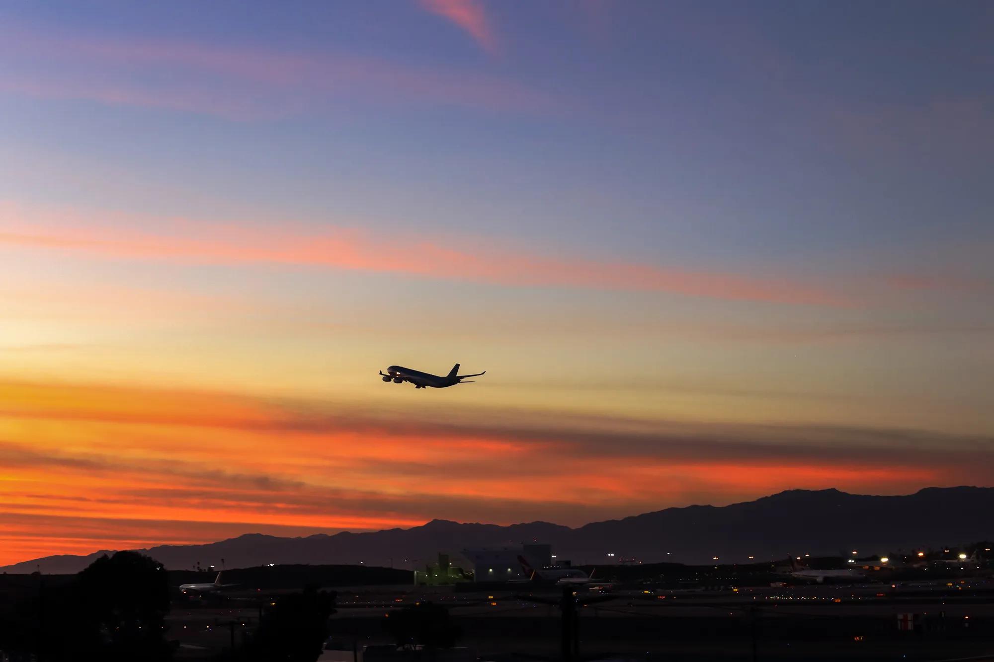 Airplane taking off at sunset above an airport, silhouetted against an orange and pink sky with distant mountains.