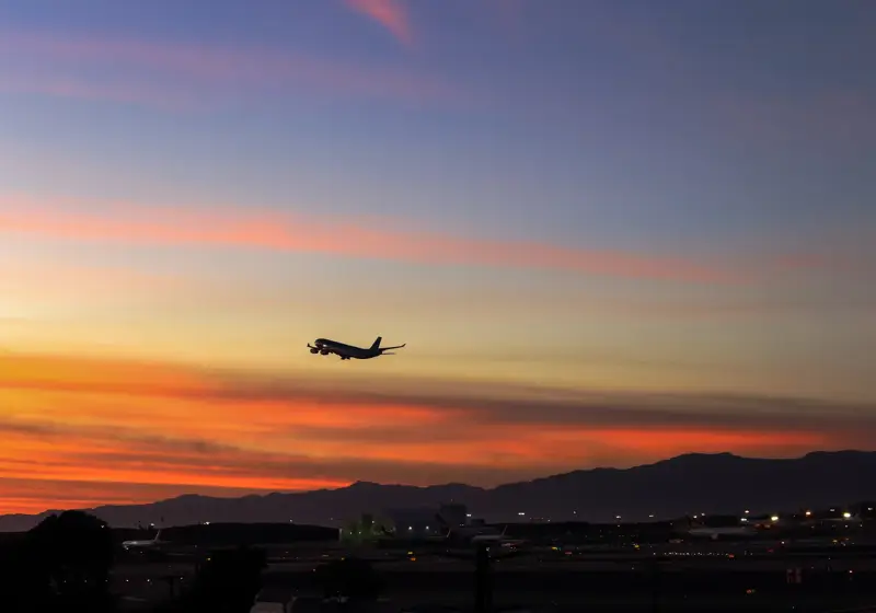 Airplane taking off at sunset above an airport, silhouetted against an orange and pink sky with distant mountains.