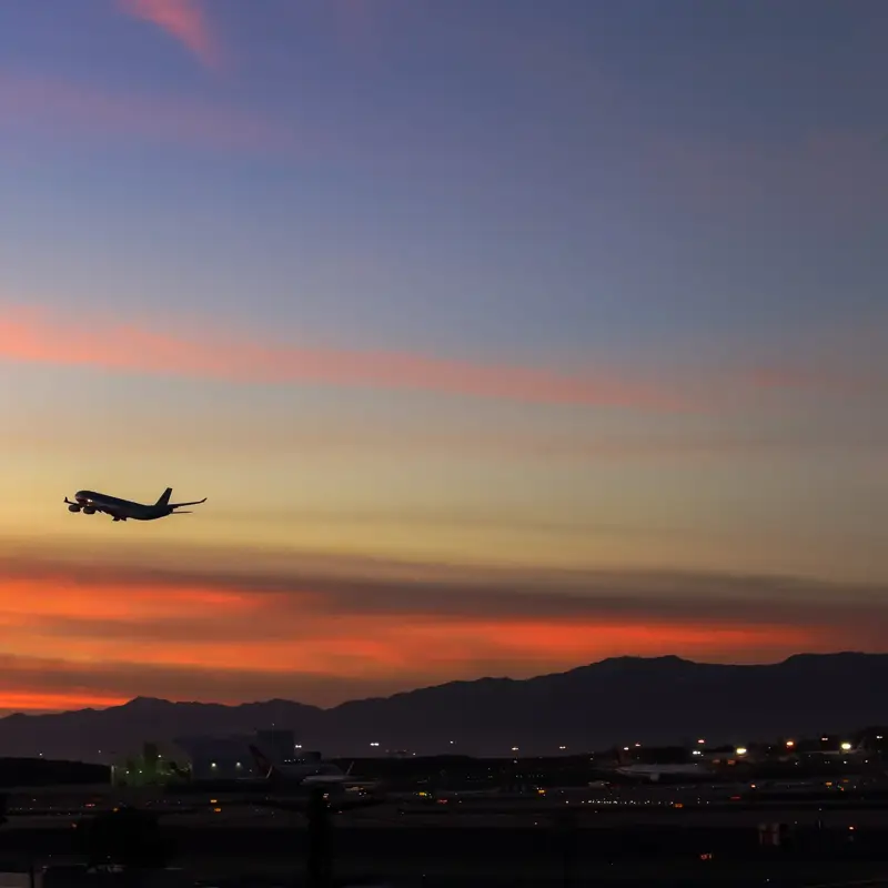 Airplane taking off at sunset above an airport, silhouetted against an orange and pink sky with distant mountains.
