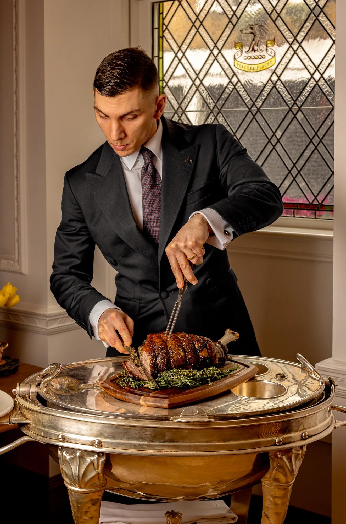 Server in a dark suit carving a roasted joint of meat on a silver trolley beside a stained-glass window.