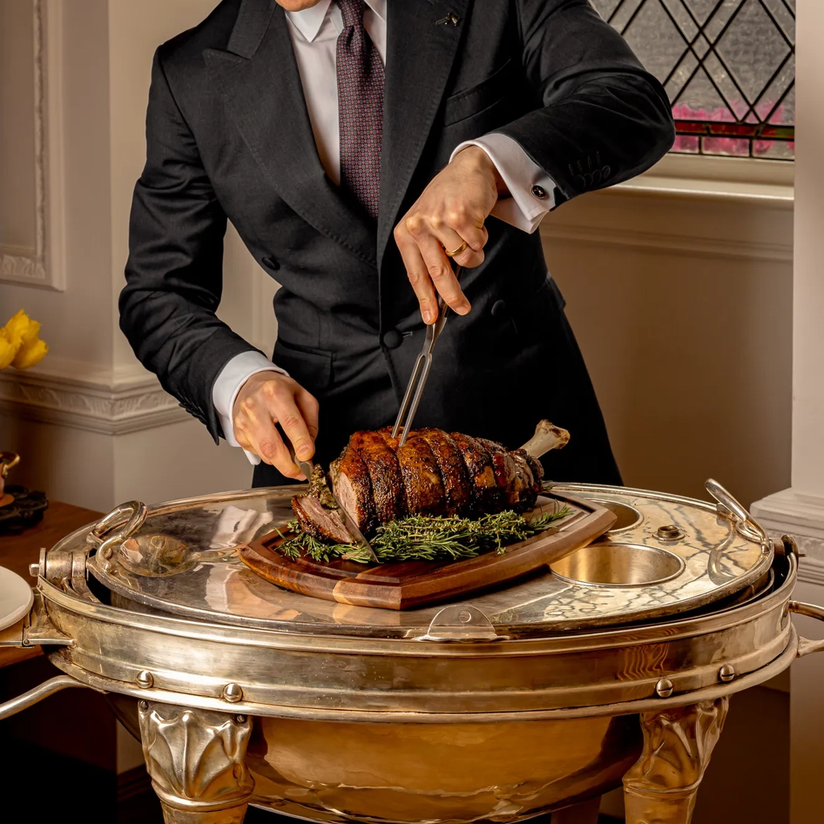 Server in a dark suit carving a roasted joint of meat on a silver trolley beside a stained-glass window.