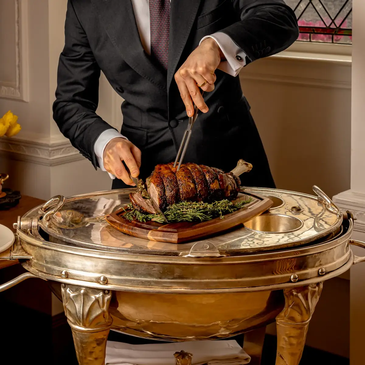 Server in a dark suit carving a roasted joint of meat on a silver trolley beside a stained-glass window.