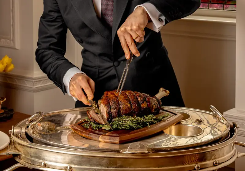 Server in a dark suit carving a roasted joint of meat on a silver trolley beside a stained-glass window.