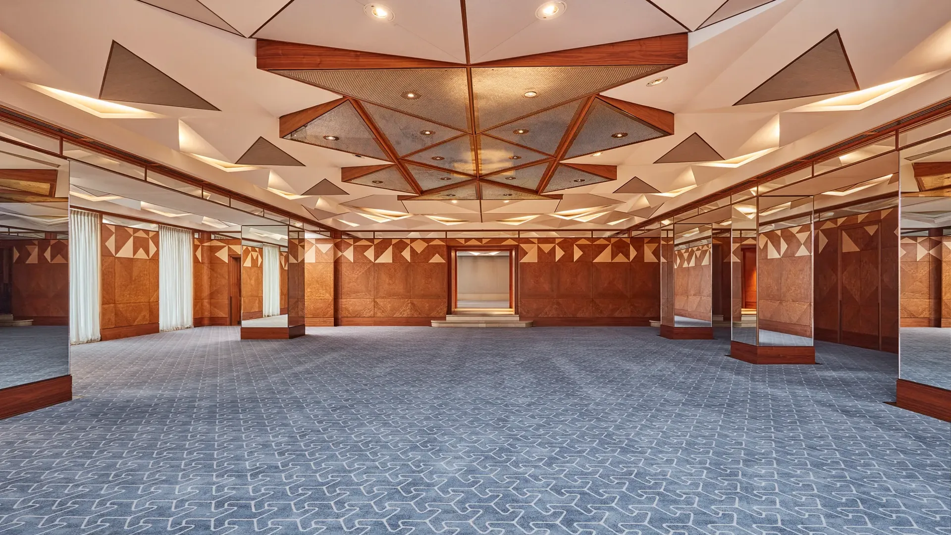 Spacious hotel ballroom with geometric ceiling design, patterned carpet, and mirrored walls for events