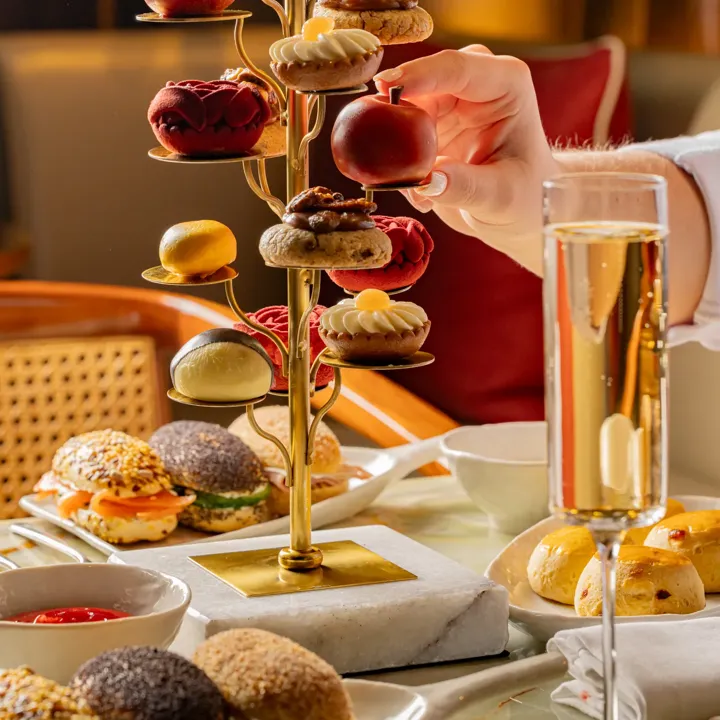 Hand holding red apple-shaped pastry from ornate afternoon tea stand with pastries, mini sliders and a champagne flute on table.