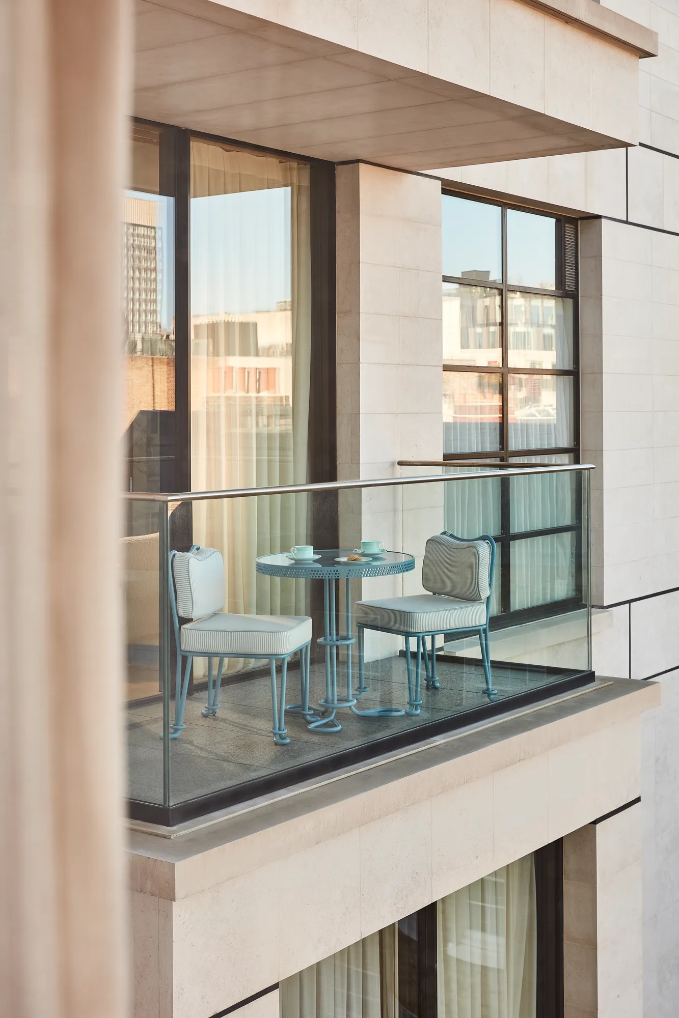 Private balcony with a glass balustrade, featuring a small blue table set with two matching chairs overlooking the city’s contemporary architecture.