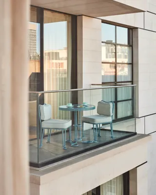 Private balcony with a glass balustrade, featuring a small blue table set with two matching chairs overlooking the city’s contemporary architecture.