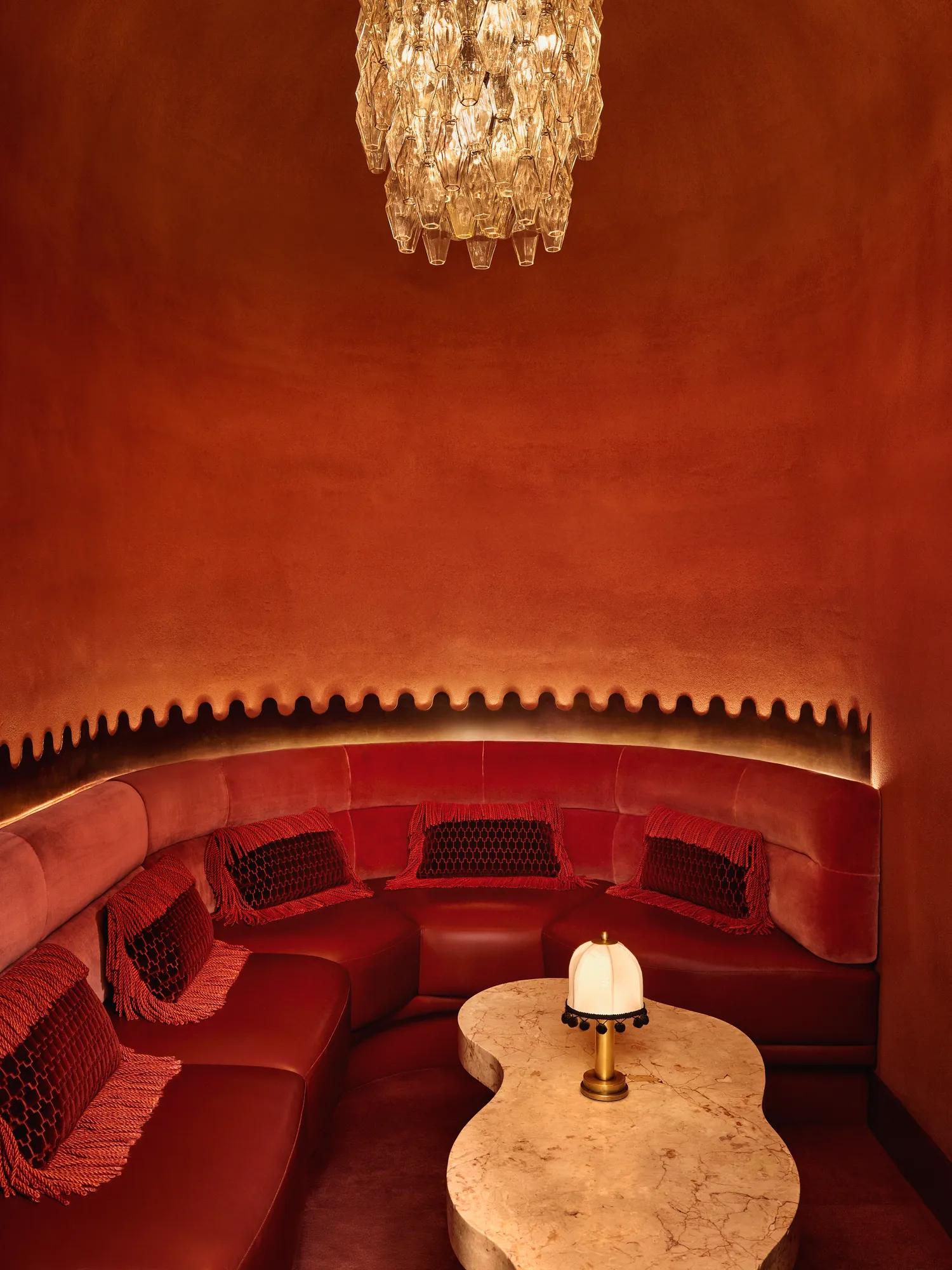 Intimate red alcove with curved velvet seating, a sculpted marble table, and a crystal chandelier overhead.