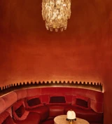 Intimate red alcove with curved velvet seating, a sculpted marble table, and a crystal chandelier overhead.
