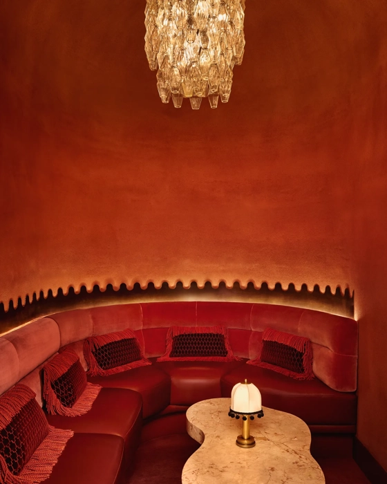 Intimate red alcove with curved velvet seating, a sculpted marble table, and a crystal chandelier overhead.