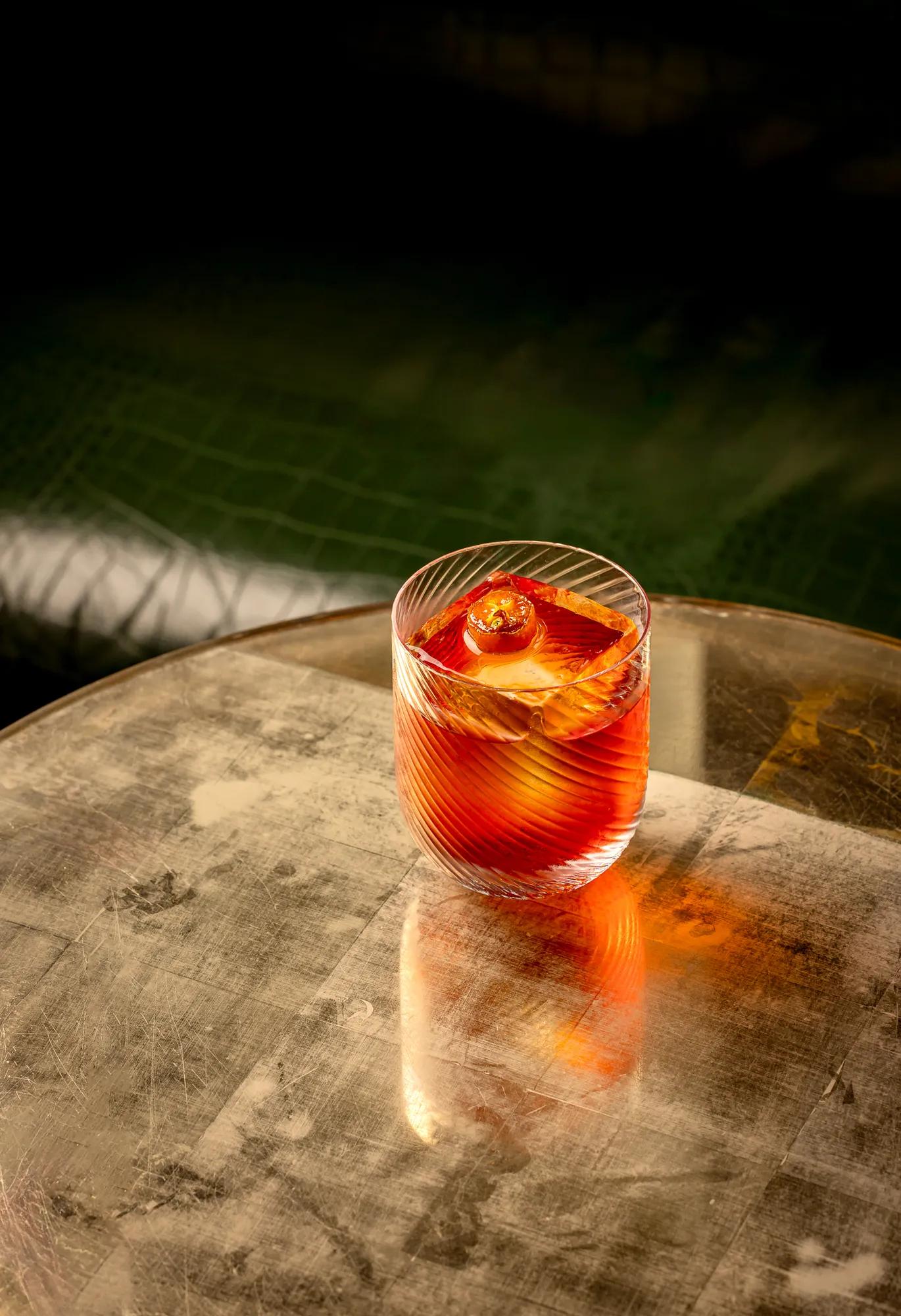 Red cocktail with orange garnish in a patterned glass, placed on a round marble table beside a softly lit pool