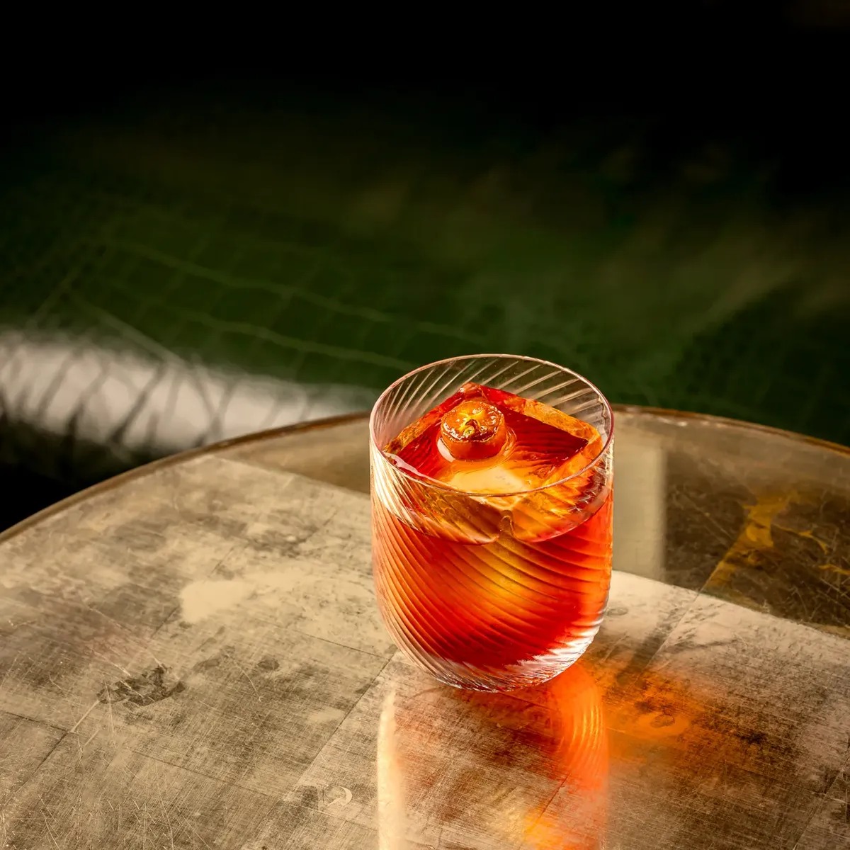 Red cocktail with orange garnish in a patterned glass, placed on a round marble table beside a softly lit pool