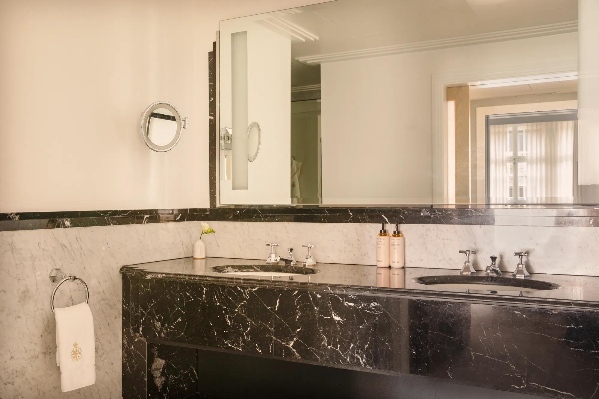 Bathroom with a black marble double vanity, large illuminated mirror, and white marble walls, viewed toward the adjoining bedroom through an open doorway.