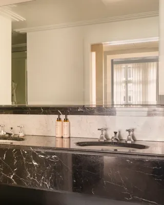 Bathroom with a black marble double vanity, large illuminated mirror, and white marble walls, viewed toward the adjoining bedroom through an open doorway.
