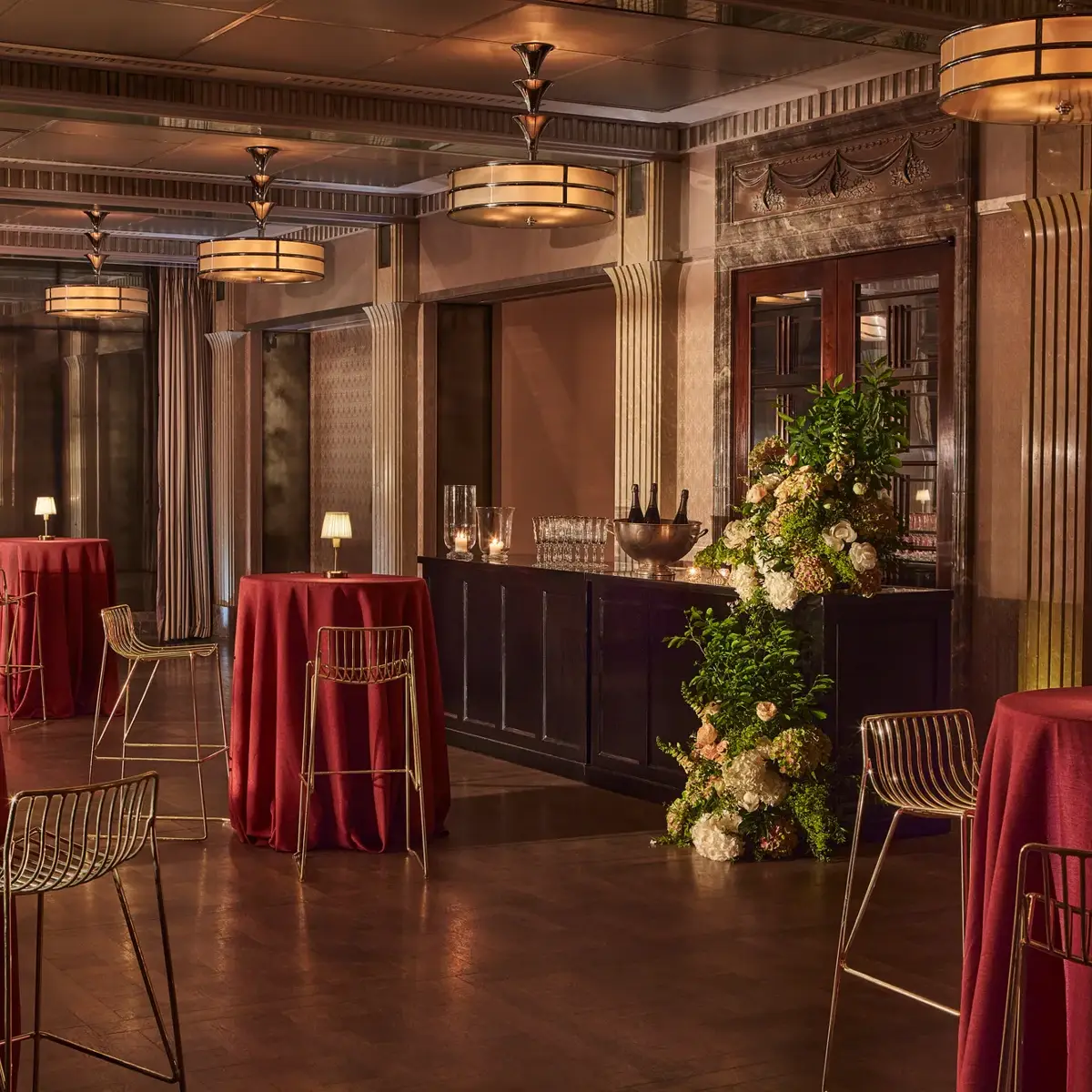 Art deco event space with red cocktail tables, gold chairs, floral display, and warm ambient lighting.