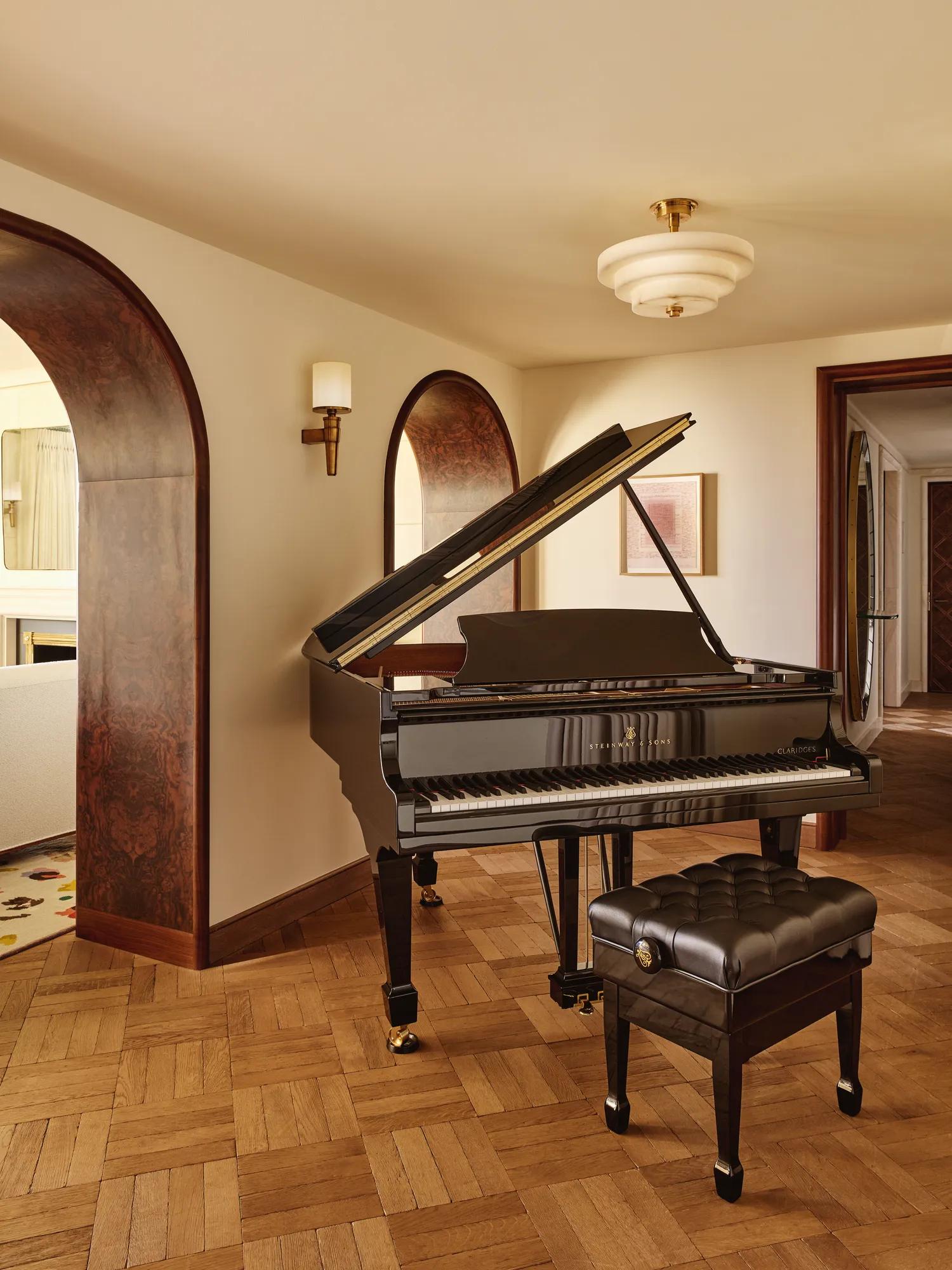 Black grand piano with a padded bench in a warm, elegant room with parquet flooring and arched doorways.