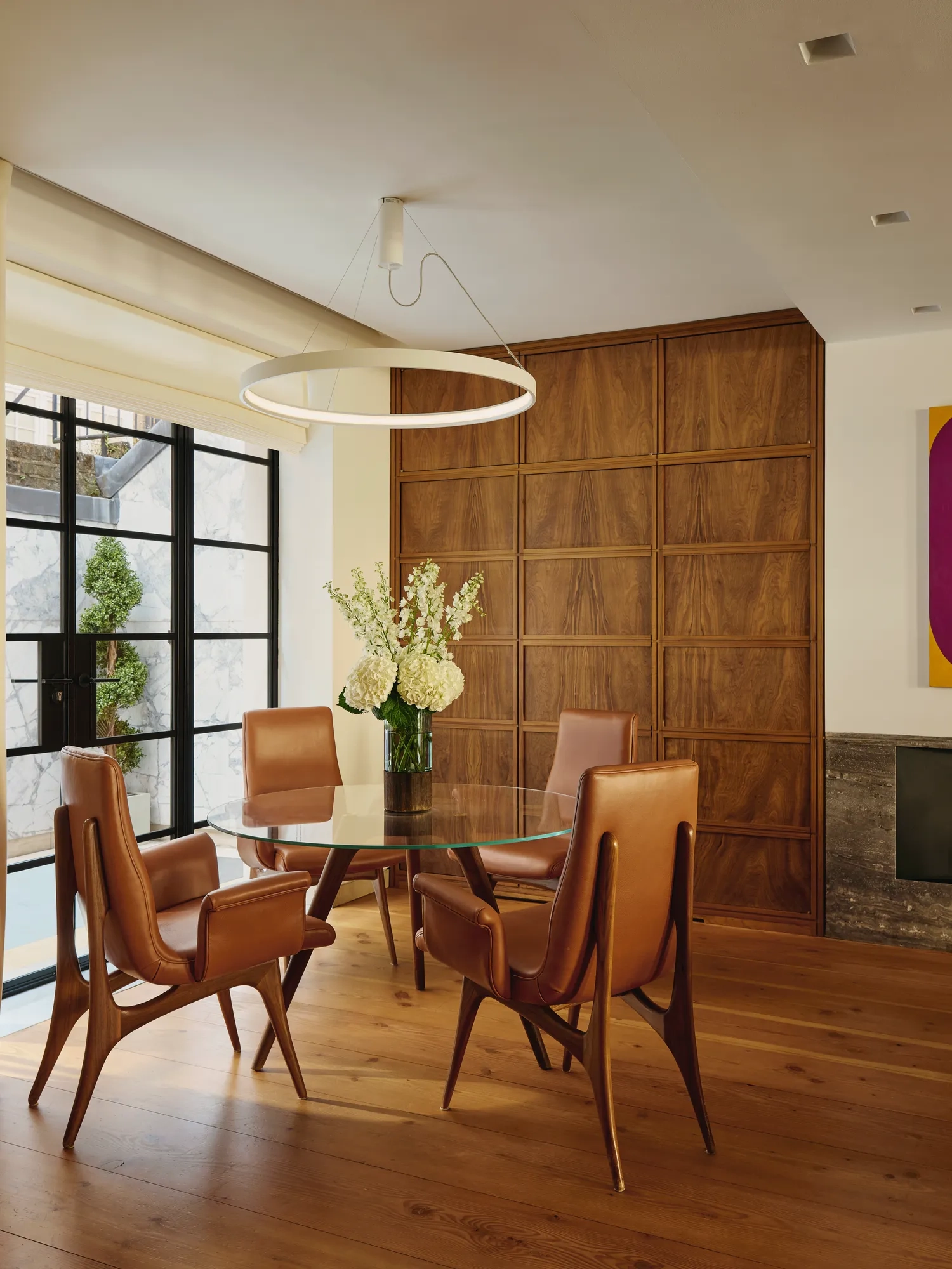 Modern dining nook with glass table, tan leather chairs, circular light, and wood-paneled wall.