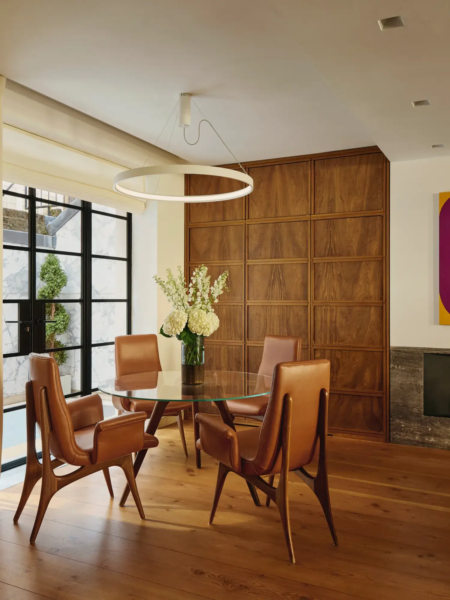 Modern dining nook with glass table, tan leather chairs, circular light, and wood-paneled wall.