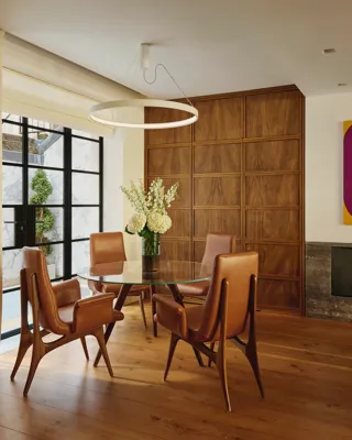 Modern dining nook with glass table, tan leather chairs, circular light, and wood-paneled wall.