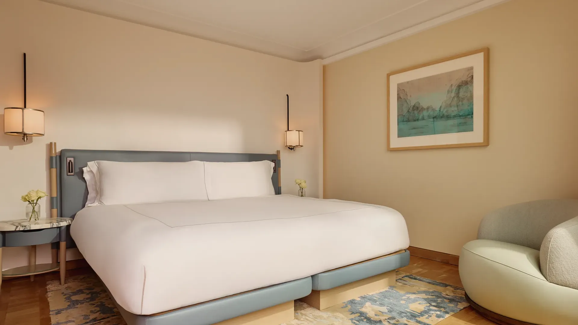 Bedroom with a king-size bed in pale blue and cream tones, bedside tables, and framed landscape artwork on the wall.